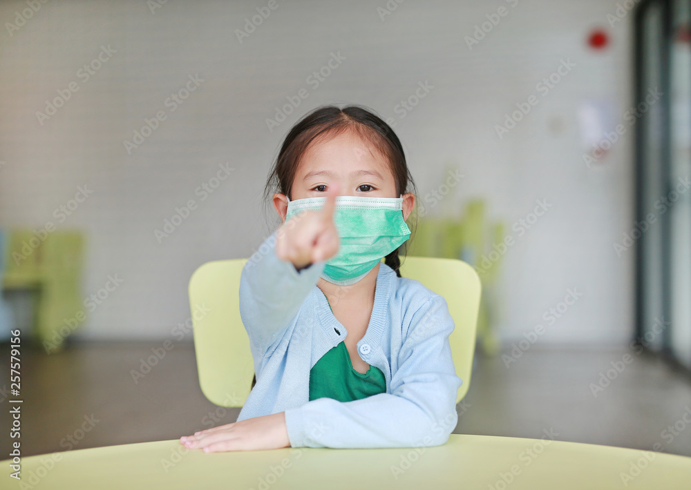 Cute little Asian child girl wearing a protective mask with pointing at you sitting on kid chair in children room. Cute little Asian child girl wearing a protective mask with pointing at you sitting on kid chair in children room.