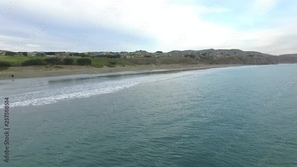 Shot of camera sweeping over the ocean heading towards the shore where there's green hills, homes, and people walking Shot of camera sweeping over the ocean heading towards the shore where there's green hills, homes, and people walking