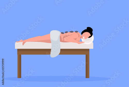 young girl client enjoying traditional hot stone massage patient woman lying on bed luxury spa and wellness center concept horizontal full length blue background