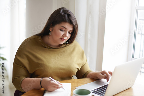 Picture of attractive young European female journalist wearng stylish earrings and knitted sweater making notes in diary while working on research for her new article, looking for information online