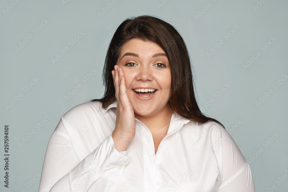 Isolated shot of plus size excited young female employee expressing astonishment and amazement, being unexpectedly promoted, exclaiming, holding hand on cheek. Positive human emotions and feelings Isolated shot of plus size excited young female employee expressing astonishment and amazement, being unexpectedly promoted, exclaiming, holding hand on cheek. Positive human emotions and feelings