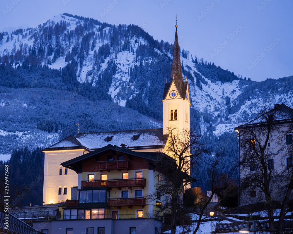 Illuminated Catholic church St. Margaretha with street, lights, mountains and forest in Kaprun Austria at blue hour