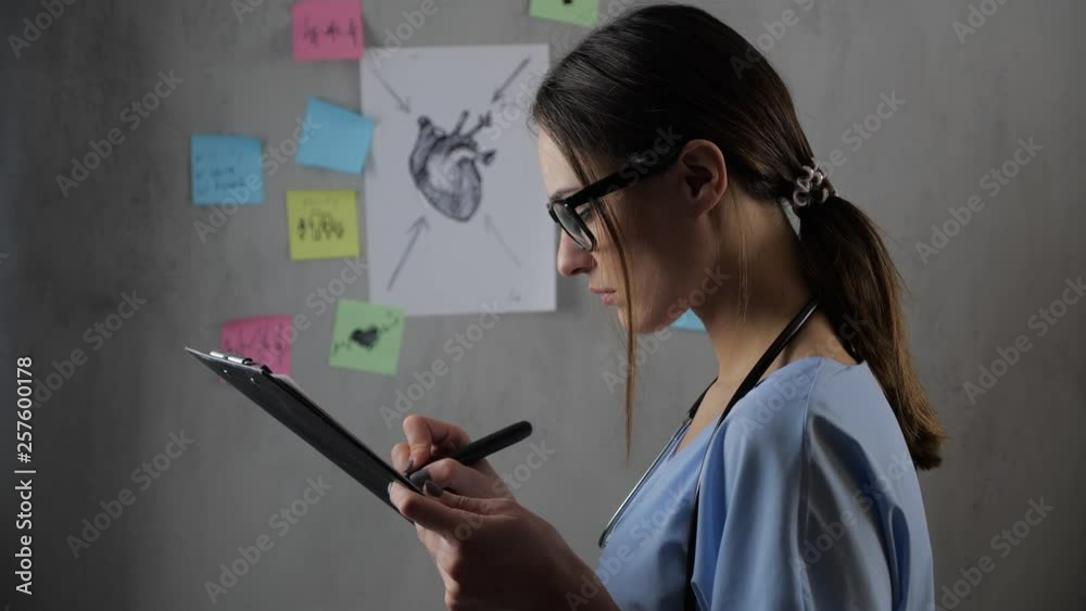 Portrait of a beautiful female doctor student with glasses preparing a presentation, against the background. Young woman draws and notes on paper. Concept: ideas, science, teacher, memory, biology Portrait of a beautiful female doctor student with glasses preparing a presentation, against the background. Young woman draws and notes on paper. Concept: ideas, science, teacher, memory, biology
