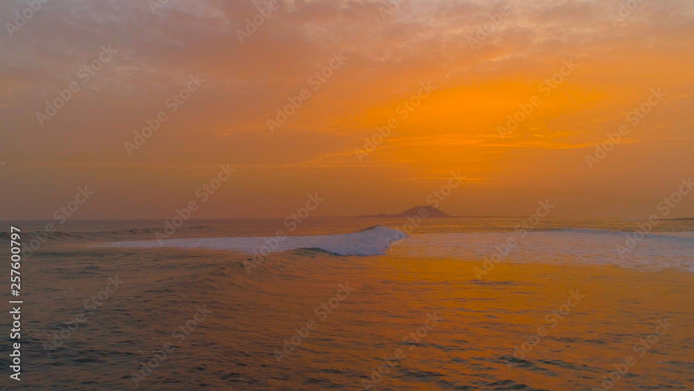 DRONE: Flying over a small ocean wave rolling towards the beach at sunrise.