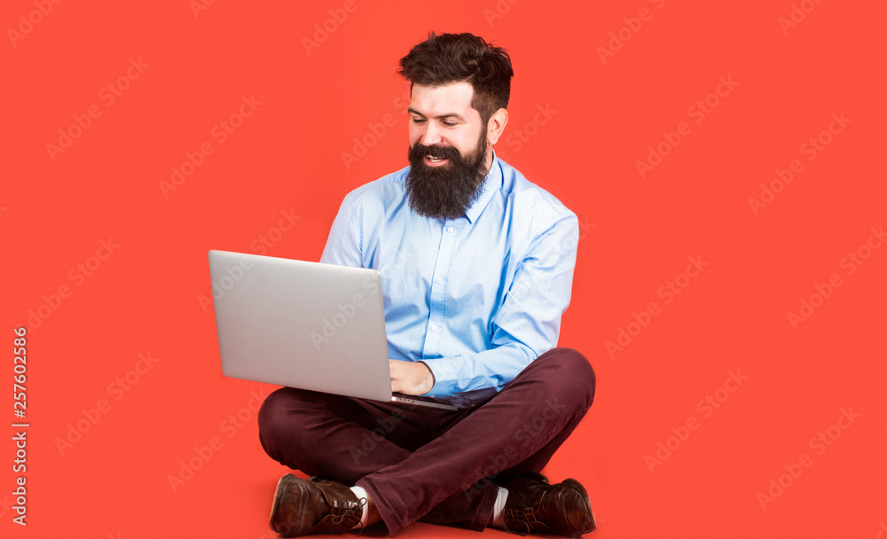 Happy young man sitting on the floor with and using laptop computer on red background. Holding laptop computer.Young businessman using his laptop, pc. Smiling handsome bearded man worker laptop.