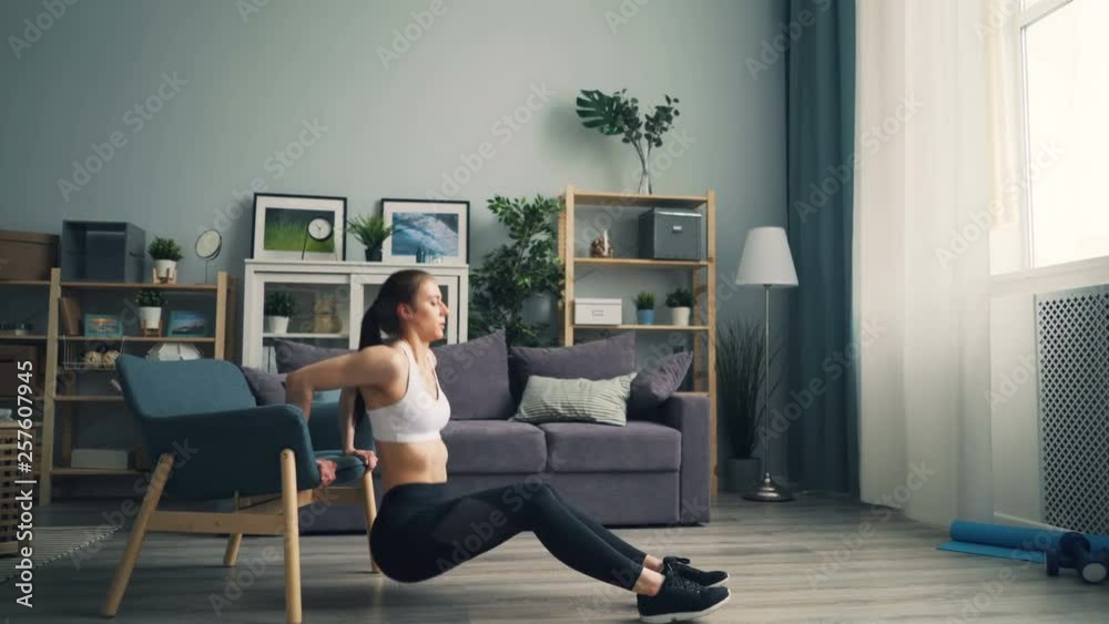 Side view of fit young woman doing pushup dips holding armchair working out in apartment wearing trendy sportswear. People, interior and training concept. Side view of fit young woman doing pushup dips holding armchair working out in apartment wearing trendy sportswear. People, interior and training concept.