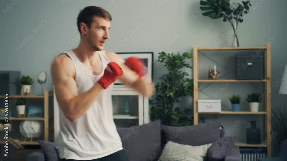 Muscular guy in sportswear is boxing alone indoors in apartment enjoying activity moving wrapped hands. Beautiful light flat is visible in background. Muscular guy in sportswear is boxing alone indoors in apartment enjoying activity moving wrapped hands. Beautiful light flat is visible in background.