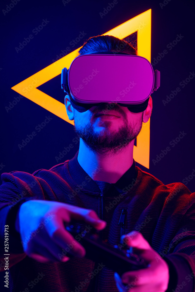 Man wearing virtual reality goggles with using game pad wireless controller. VR headset videogame in 80's wave and retrowave glowing triangle futuristic aesthetics. foto de Stock | Adobe Stock