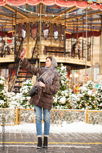 Young woman in mink fur coat taking pictures of himself on a cell phone in the center of the Christmas courtyard