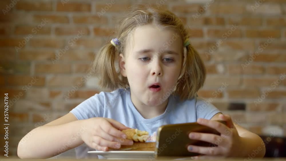 Cute little girl eating sandwich while watchine funny videos online on smartphone. Cute little girl eating sandwich while watchine funny videos online on smartphone.