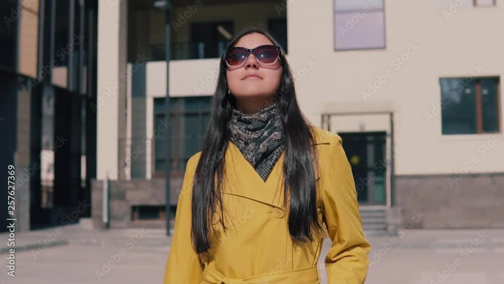 stylish woman in a yellow raincoat with long dark hair goes with luggage removes glasses looking around and smiling stylish woman in a yellow raincoat with long dark hair goes with luggage removes glasses looking around and smiling