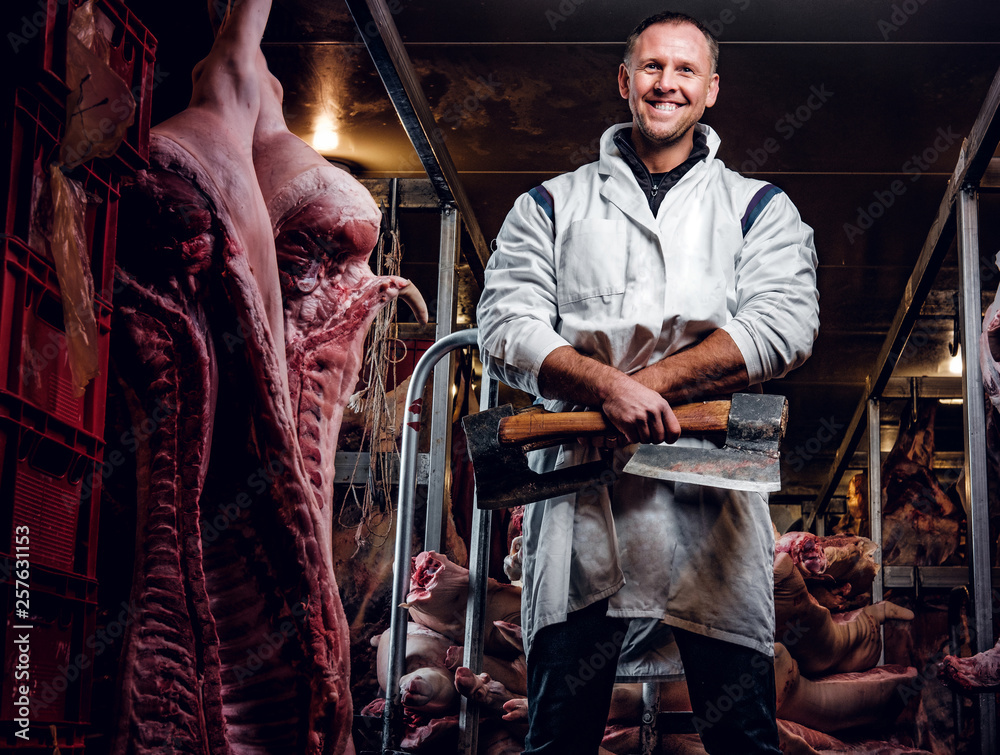 Cheerful butcher in workwear posing with two axes in a refrigerated warehouse in the midst of meat carcasses Cheerful butcher in workwear posing with two axes in a refrigerated warehouse in the midst of meat carcasses