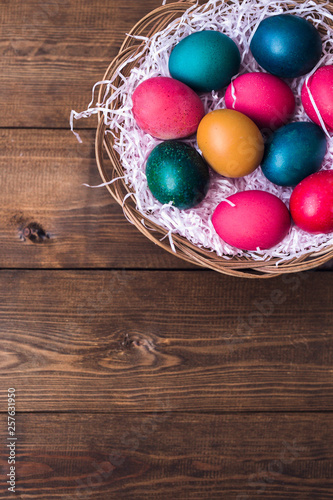 close up of colored eggs in wicker basket over brown wooden boards background