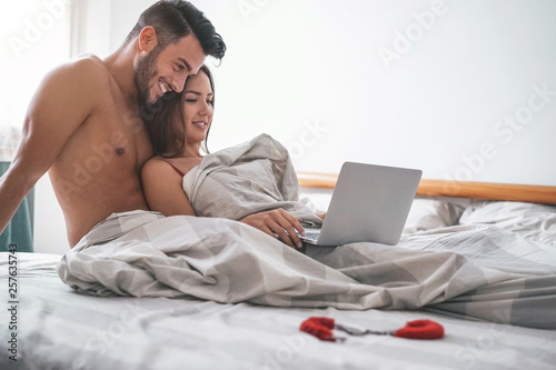 Obraz na plátně Happy couple watching on computer while lying on bed under blanket - Young lover
