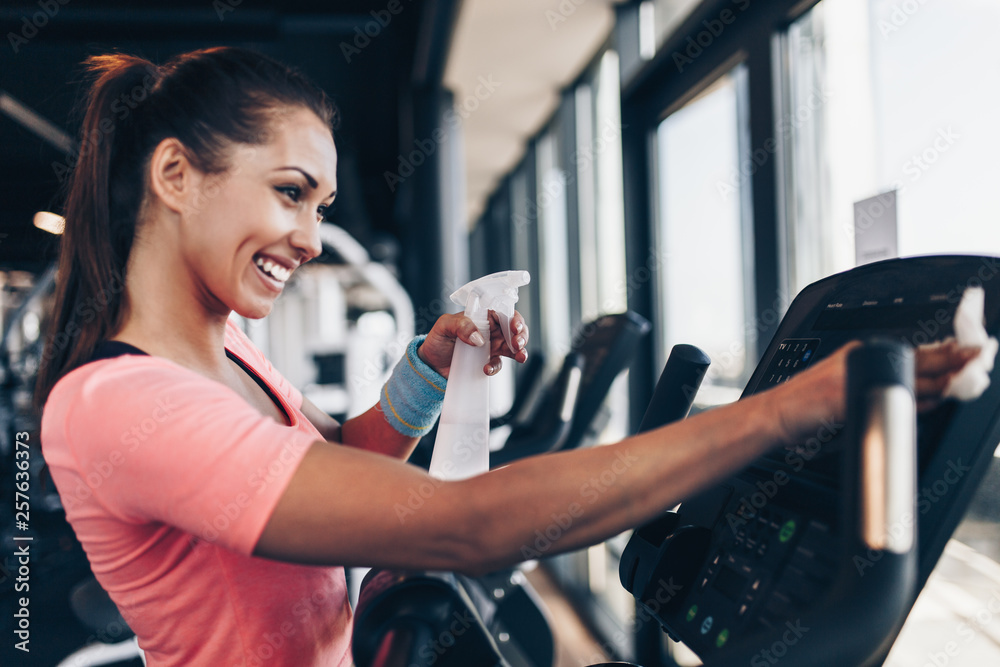 Young happy and smiling woman cleaning and weeping expensive fitness gym  equipment with sprayer and cloth. Stock Photo | Adobe Stock