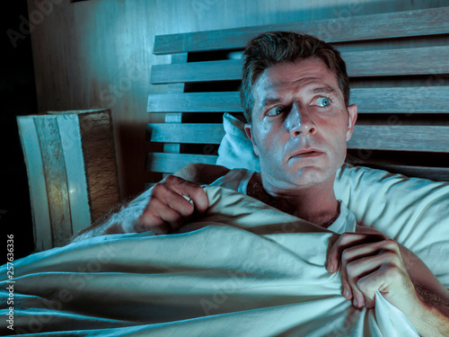 sleepless young man lying in bed stressed and scared suffering nightmare and horror bad dream grabbing duvet frightened and paranoid in sleeping disorder