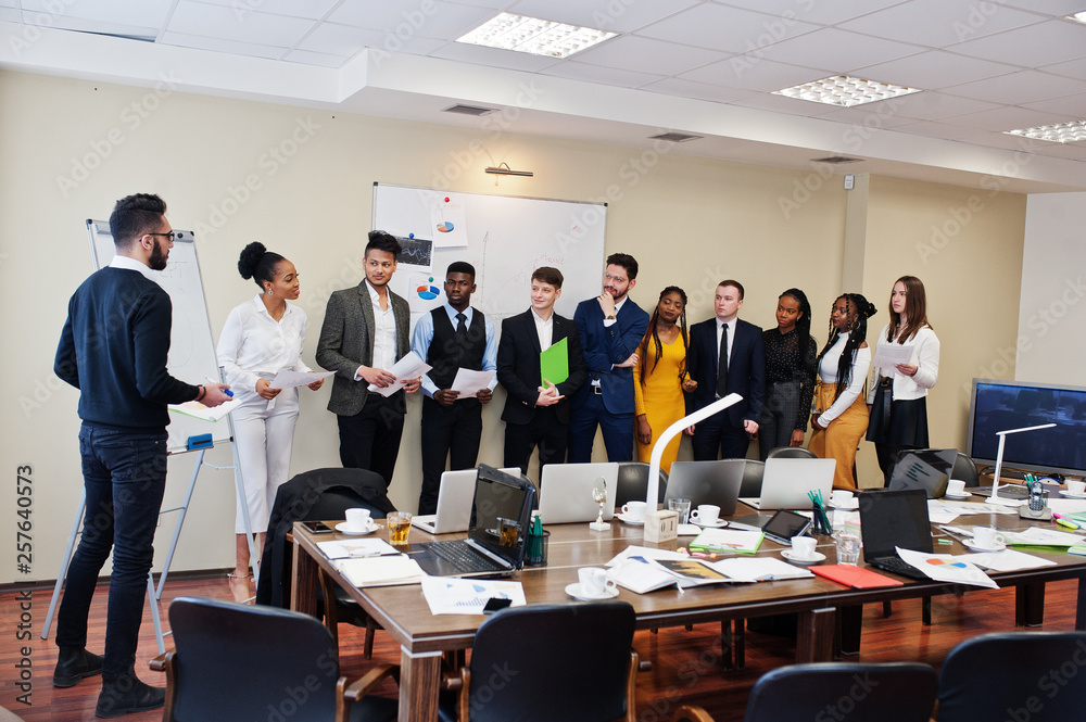 Mixed race business coach presenting report standing near whiteboard pointing on sales statistic shown on diagram and chart teach diverse company members gathered together in conference room. Mixed race business coach presenting report standing near whiteboard pointing on sales statistic shown on diagram and chart teach diverse company members gathered together in conference room.