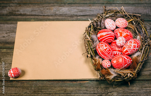 Easter eggs with white pattern inside bird nest on right side of old sheet of paper on wood board. Top view horizontal background.