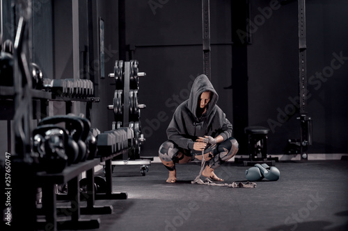 Young Caucasian woman with hoodie crouching barefoot and putting bandages on hand. Next to her boxing gloves. Night workout concept.