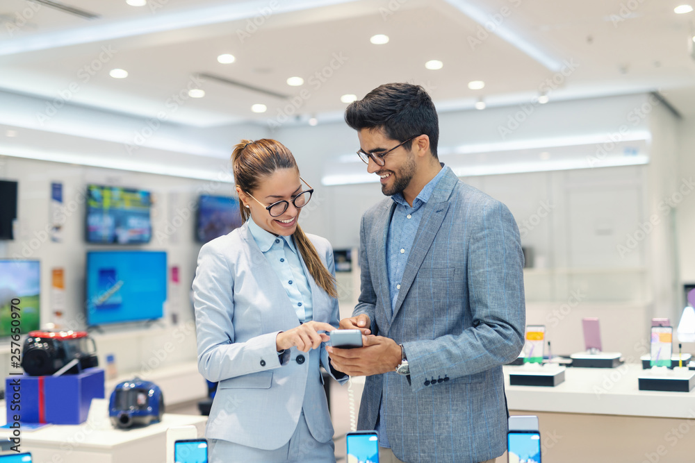 Smiling multicultural couple in formal wear trying out new smart phone while standing in tech store. Smiling multicultural couple in formal wear trying out new smart phone while standing in tech store.