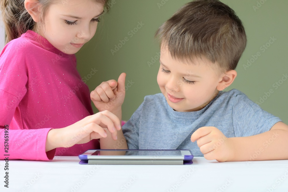 White caucasian toddler boy and beautiful girl smiling and playing together on tablet games. Portrait of a happiness little brother and sister. Happy Children play a computer game on the tablet. White caucasian toddler boy and beautiful girl smiling and playing together on tablet games. Portrait of a happiness little brother and sister. Happy Children play a computer game on the tablet.