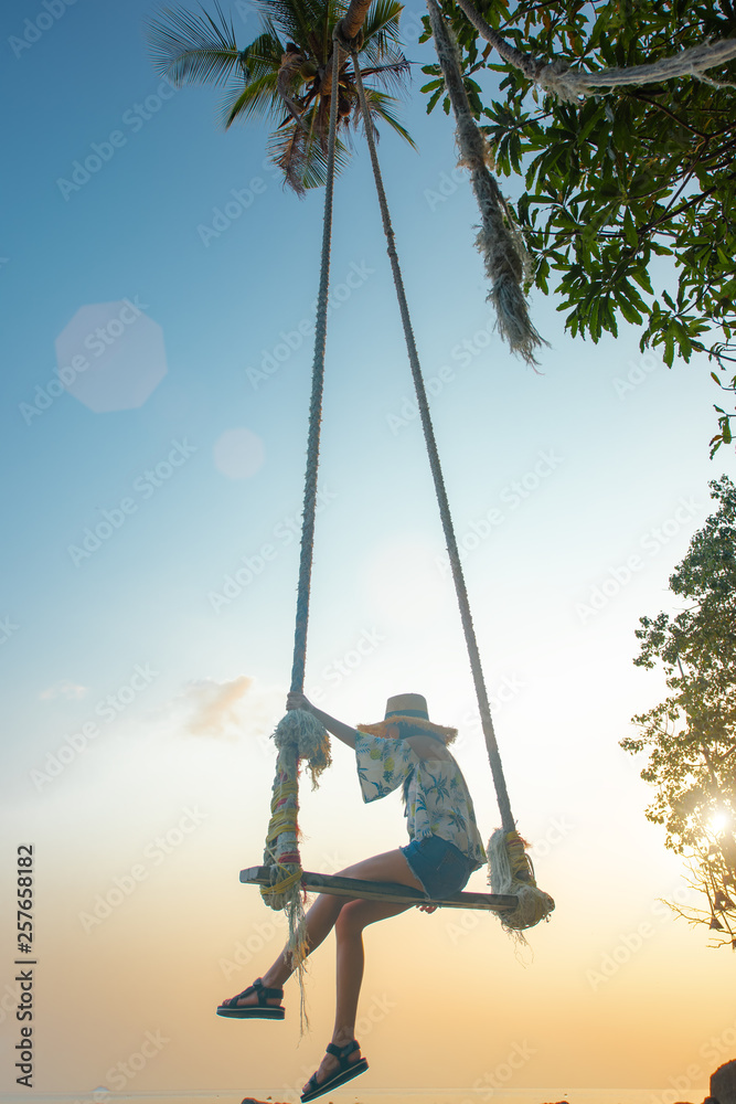 young woman sitting on sway the wooden swing under coconut palm tree and shadow of the trees over the sea beach, sunbeam and sunset light feel comfortable and most relaxation .