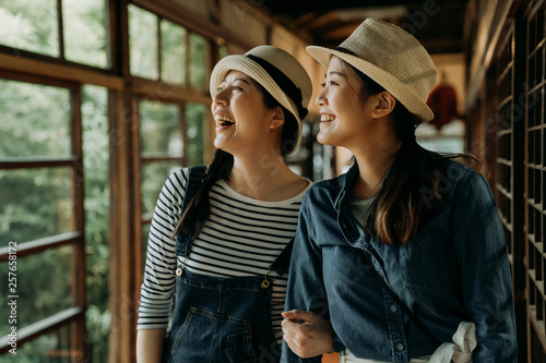 Fototapeta dwie azjatyckie podróżniczki w słomkowych kapeluszach uśmiechnięte śmiejące się zwiedzanie wiosenny widok na ogród podczas spaceru po japońskim drewnianym chodniku. szczęśliwe wesołe kobiety relaksujące podróże w tokio japonia styl życia.