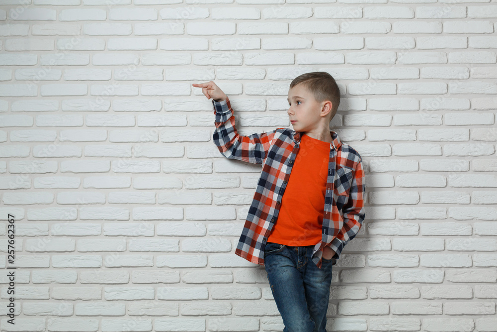 Cute stylish boy near white brick wall . boy points Finger at something isolated on white background. Portrait of a caucasian boy in jeans and a shirt in a cell. boy shows something with a finger. Cute stylish boy near white brick wall . boy points Finger at something isolated on white background. Portrait of a caucasian boy in jeans and a shirt in a cell. boy shows something with a finger.
