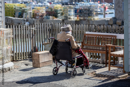 An elderly woman sits in her wheelchair warmly dressed outside in a New England sea side setting.