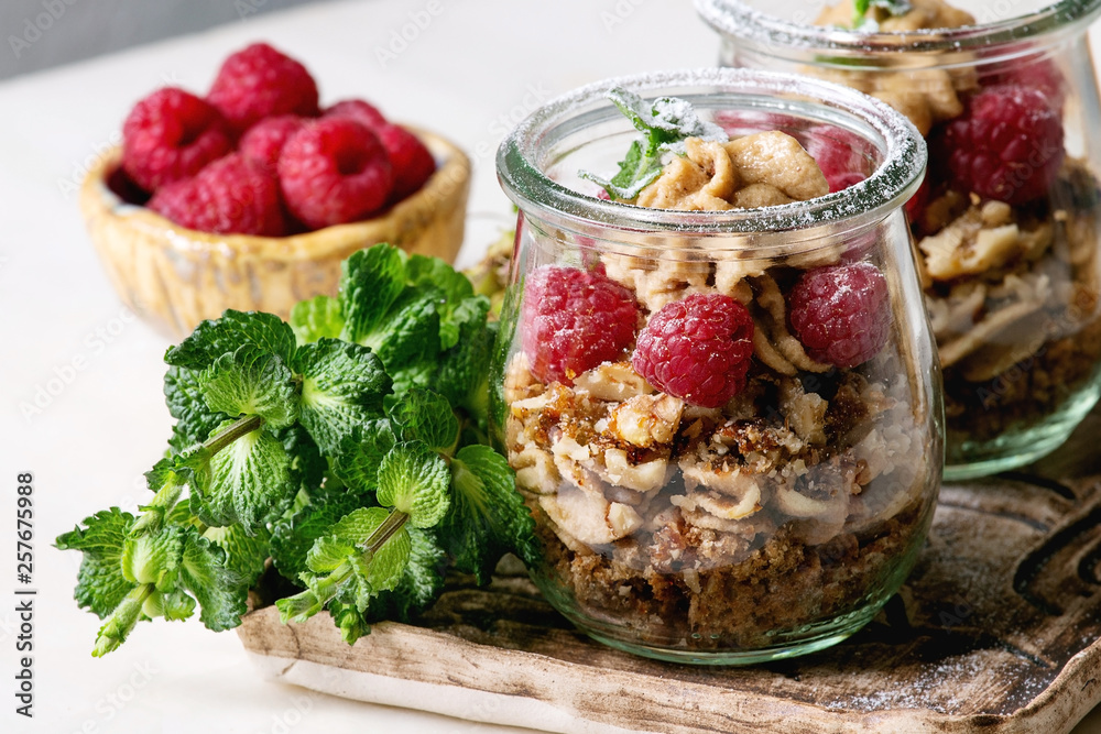 Layered dessert in jars. Biscuit, coffee cream, nuts, raspberries, fresh mint. Served on ceramic tray with berries and greens over white marble table. Layered dessert in jars. Biscuit, coffee cream, nuts, raspberries, fresh mint. Served on ceramic tray with berries and greens over white marble table.