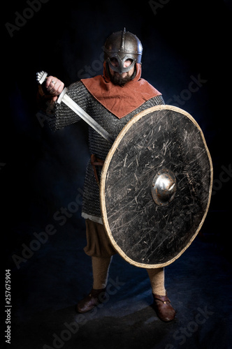 Young bearded man in a Viking-era helmet covering his face standind and holding a sword and shield