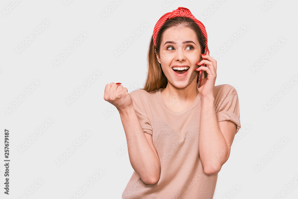 Young beautiful woman in beige t-shirt, red headband, smiling and talking on smart phone to her friend, looking cheerful and happy, posing on white studio background. Real human emotions. Young beautiful woman in beige t-shirt, red headband, smiling and talking on smart phone to her friend, looking cheerful and happy, posing on white studio background. Real human emotions.