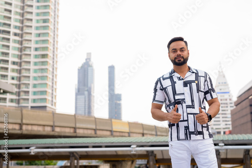 Young bearded Indian man giving thumbs up against view of sky train station
