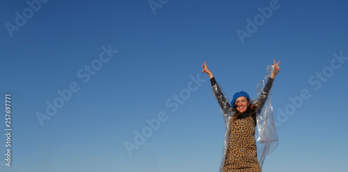 girl with her hands up looks at the camera and smiles. Dressed in a dress, a transparent raincoat and a blue hat. Against the blue sky. Day. Spring, summer or autumn. Horizontally