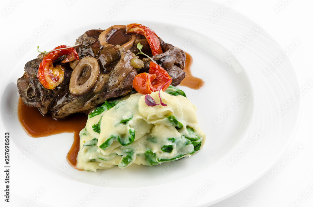 Braised beef on the bone with dried tomatoes and mashed potatoes with spinach and feta cheese on a plate on a white background Braised beef on the bone with dried tomatoes and mashed potatoes with spinach and feta cheese on a plate on a white background