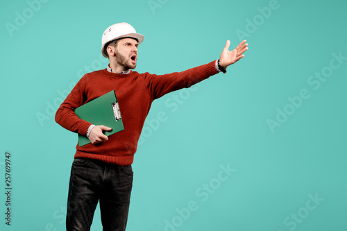 Unhappy young handsome bearded engineer or constructor man in casual outfit holding green clipboard over cyan background. Architecture and Renovation concept.