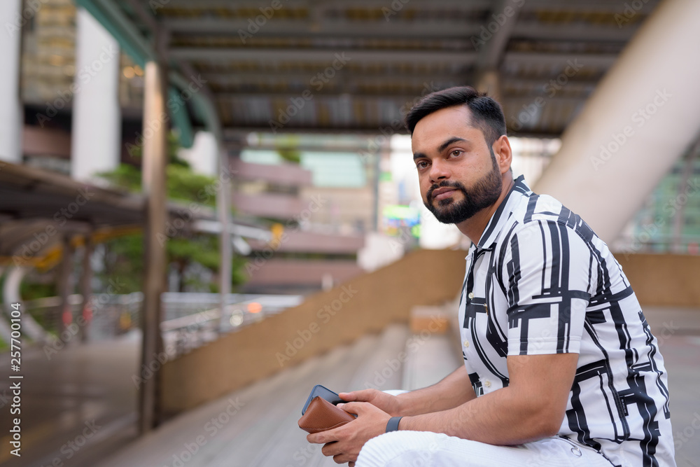 Obraz premium Young bearded Indian man thinking with phone on staircase at the city