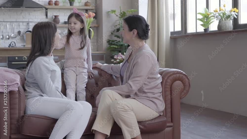 Grandmother, mother and little daughter spend time together sitting on the sofa in modern apartment. The girl standing on the cout, mom and granny looking at her smiling. Concept of generation. Grandmother, mother and little daughter spend time together sitting on the sofa in modern apartment. The girl standing on the cout, mom and granny looking at her smiling. Concept of generation.