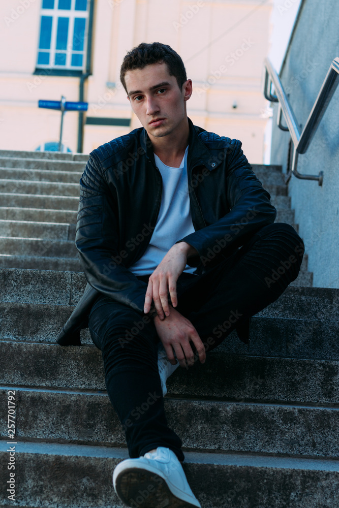 Sexy man posing sits on the steps near railing. Handsome young man in stylish black clothes and white shoes posing near a railing Sexy man posing sits on the steps near railing. Handsome young man in stylish black clothes and white shoes posing near a railing