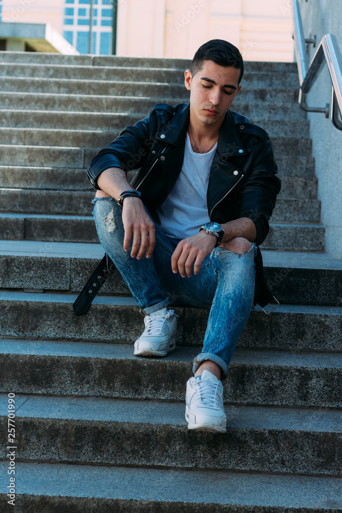 Sexy man posing sits on the steps near railing. Handsome young man in stylish black clothes and white shoes watch on hand Sexy man posing sits on the steps near railing. Handsome young man in stylish black clothes and white shoes watch on hand