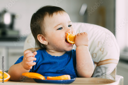 little girl sitting in baby chair, eating an orange