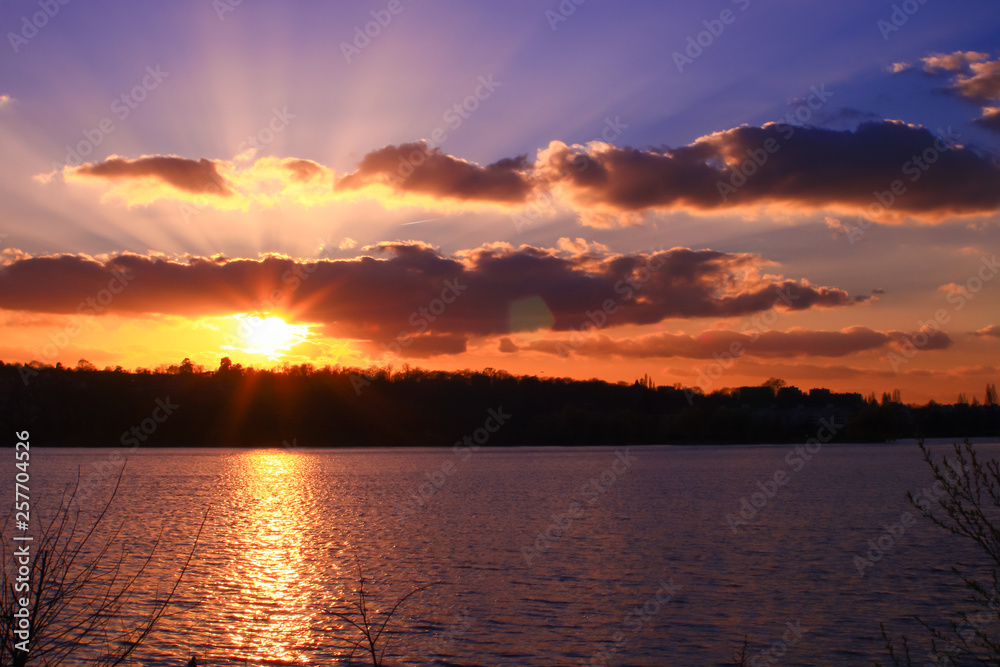 Sunset reflected on the water with a forest in the background. Sun rays through pretty orange colored clouds in a blue sky