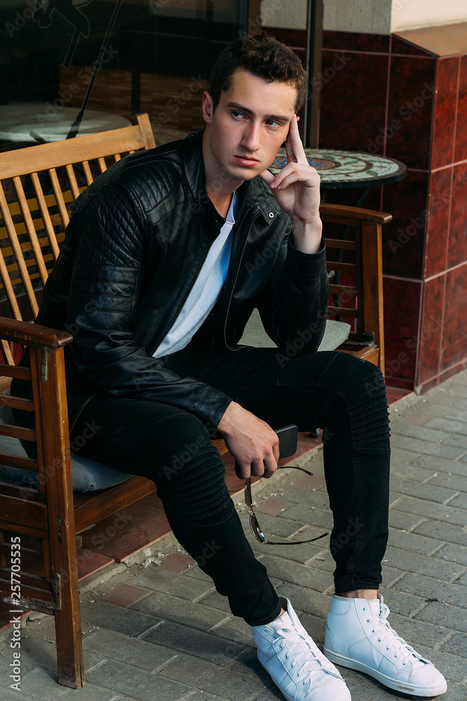 attractive guy sitting on a bench in a cafe, on the street, white sneakers, white T-shirt, black leather jacket and black jeans, pants. model. waiting for a meeting. attractive guy sitting on a bench in a cafe, on the street, white sneakers, white T-shirt, black leather jacket and black jeans, pants. model. waiting for a meeting.