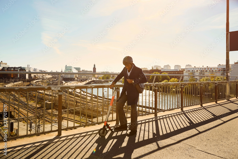 Full length of young man standing with electric push scooter by railing on bridge in city Full length of young man standing with electric push scooter by railing on bridge in city