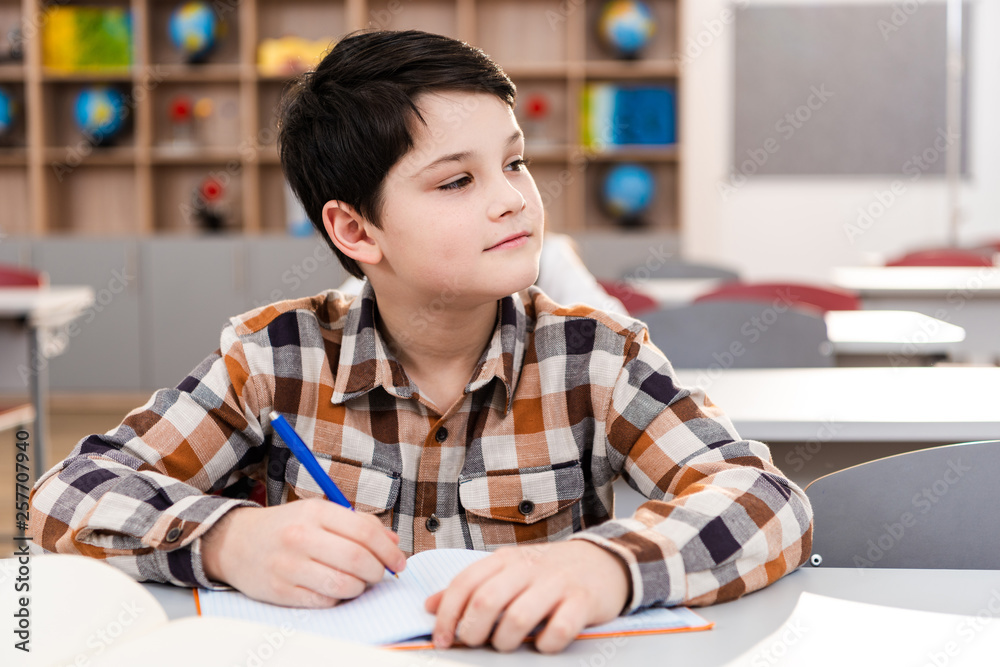 Pensive brunette pupil in checkered shirt writing in notebook during lesson Pensive brunette pupil in checkered shirt writing in notebook during lesson