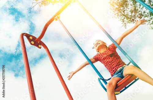 Happy smiling little girl swinging on the swing wide opened an arms with blue sky background