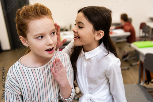Two cute schoolgirls smiling and sharing gossips in classroom during brake