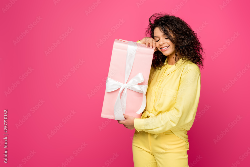 happy curly african american woman holding pink gift box with white satin ribbon isolated on crimson happy curly african american woman holding pink gift box with white satin ribbon isolated on crimson