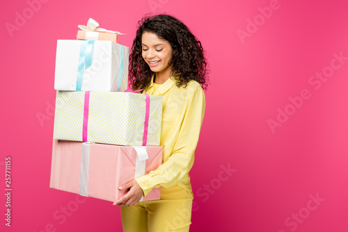cheerful curly african american woman holding gift boxes isolated on crimson
