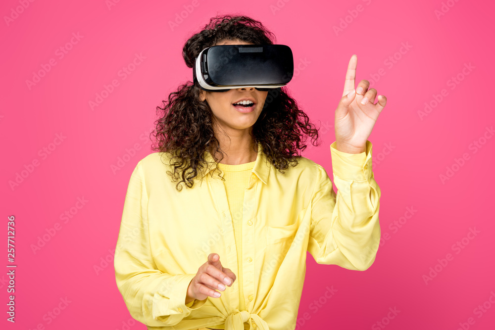 surprised curly african american woman wearing virtual reality headset and pointing with fingers isolated on crimson